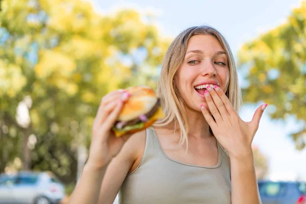 若い金髪の女性は驚きとショックの表情で屋外でハンバーガーを保持しています — ストック写真