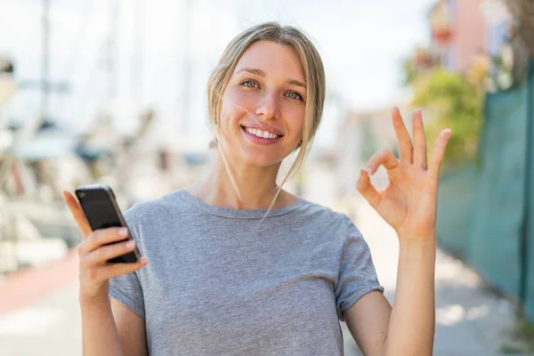 Young Blonde Woman Using Mobile Phone Outdoors Showing Sign Fingers — Stock Photo, Image