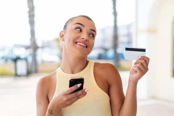 Young pretty woman at outdoors buying with the mobile with a credit card while thinking