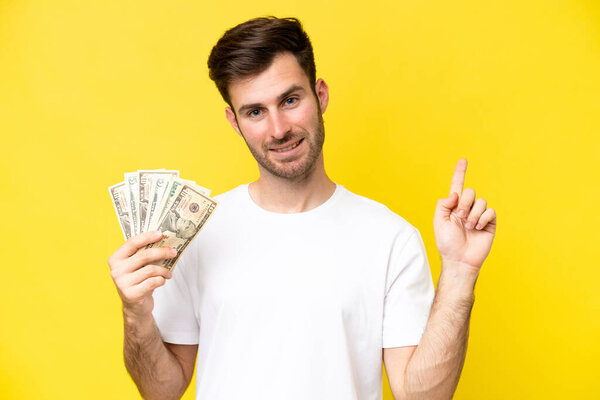 Young caucasian man taking a lot of money isolated on yellow background showing and lifting a finger in sign of the best