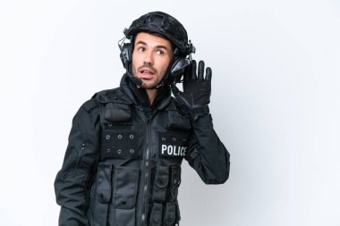 Beyaz arka planda izole edilmiş genç beyaz SWAT adamı elini kulağına koyarak bir şeyler dinliyor.