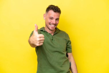 Beyaz tenli yakışıklı adam sarı arka planda izole edildi. Başparmakları havada, çünkü iyi bir şey oldu.