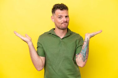 Sarı arka planda izole edilmiş beyaz, yakışıklı genç bir adam ellerini kaldırırken şüphe duyuyor.
