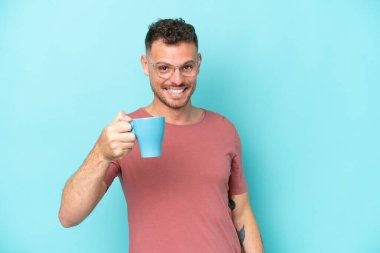 Kafkasyalı genç bir adam elinde bir fincan kahve tutarken mavi arka planda mutlu bir ifadeyle izole edilmiş.
