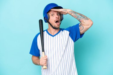 Kasklı beyzbol oyuncusu ve beyzbol sopası mavi arka planda izole edilmiş. Yan tarafa bakarken sürpriz jest yapıyor.