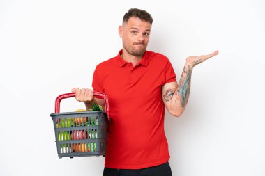 Beyaz arka planda izole edilmiş yiyecek dolu bir sepet tutan genç beyaz bir adam şüphe içinde el kaldırırken...