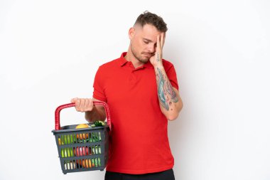 Beyaz arka planda izole edilmiş, başağrısı olan, yiyecek dolu bir alışveriş sepeti tutan genç beyaz bir adam.