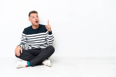 Young man sitting on the floor isolated on white background thinking an idea pointing the finger up