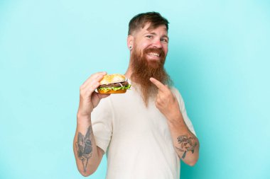 Kızıl saçlı, uzun sakallı bir adam elinde bir burger tutuyor mavi arka planda izole edilmiş ve onu işaret ediyor.