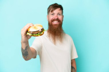 Uzun sakallı kızıl saçlı adam elinde hamburgerle mavi arka planda mutlu bir ifadeyle izole edilmiş.