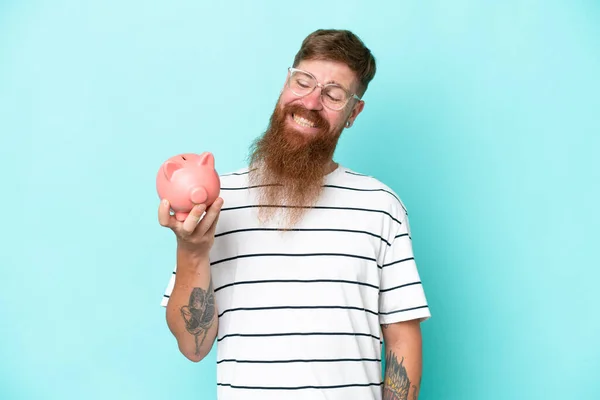 Redhead man with long beard holding a piggybank isolated on blue background with happy expression