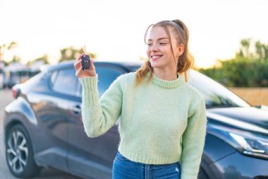 Genç ve güzel bir kız dışarıda arabanın anahtarlarını tutarken mutlu bir ifadeyle.