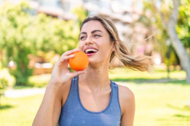 Açık havada genç sarışın bir kadın elinde mutlu bir ifadeyle bir portakal tutuyor.