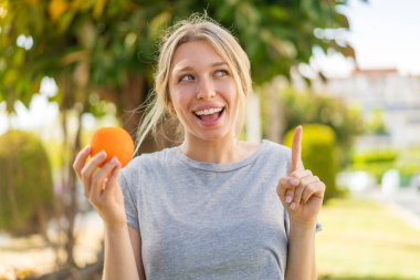 Açık havada portakal tutan genç sarışın kadın parmağını kaldırarak çözümü anlamaya çalışıyor.