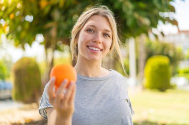 Açık havada elinde portakal tutan genç sarışın kadın. Mutlu bir ifadeyle.