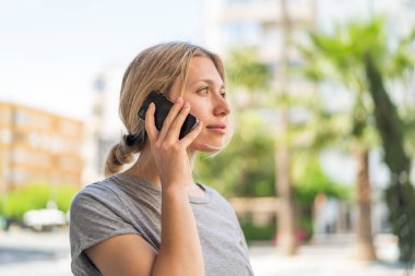 Açık havada genç sarışın bir kadın cep telefonuyla biriyle sohbet ediyor.