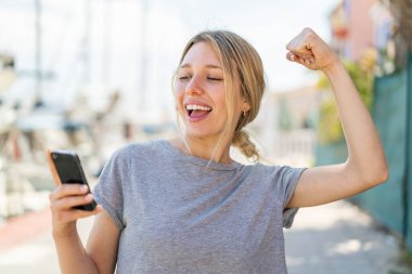 Açık havada cep telefonu kullanan genç sarışın kadın zaferi kutluyor.