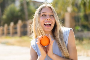 Açık havada genç sarışın bir kadın elinde turuncu bir suratla.