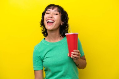 Young Argentinian woman holding a soda isolated on yellow background laughing