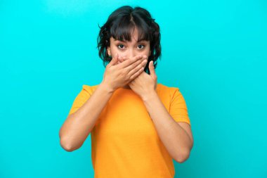 Arjantinli genç bir kadın mavi arka planda izole edilmiş, ağzı elleriyle kapatıyor.