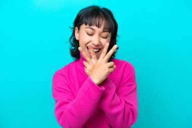 Genç Arjantinli kadın mavi arka planda izole edilmiş gülümsüyor ve zafer işareti gösteriyor.