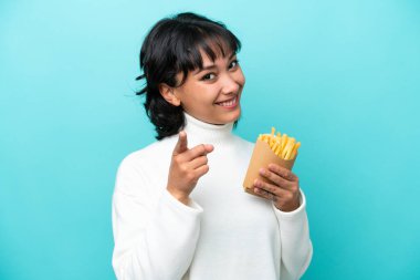 Elinde kızarmış cipsler tutan genç Arjantinli kadın mavi arka planda izole edilmiş mutlu bir ifadeyle ön tarafı işaret ediyor.