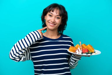 Elinde waffle tutan genç Arjantinli kadın mavi arka planda izole edilmiş bir şekilde baş parmak işareti yapıyor.