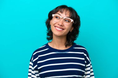 Genç Arjantinli kadın mavi arka planda izole edilmiş, mutlu bir ifadeyle gözlüklü.
