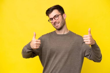 Sarı arka planda izole edilmiş genç beyaz bir adam baş parmak hareketi yapıyor.