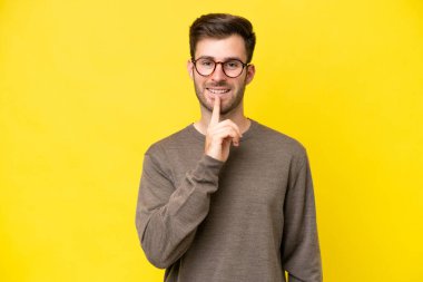 Sarı arka planda izole edilmiş genç beyaz bir adam sessizlik işareti gösteriyor. Parmağını ağzına sokuyor.
