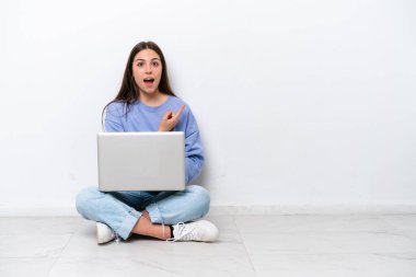 Young caucasian woman with laptop sitting on the floor isolated on white background surprised and pointing side