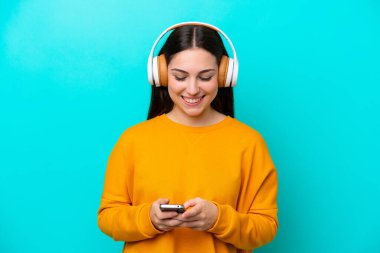 Genç kız mavi arka planda izole edilmiş müzik dinliyor ve cep telefonu arıyor.