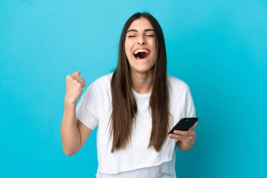Mavi arka planda izole edilmiş genç beyaz kadın cep telefonu kullanıyor ve zafer jesti yapıyor.
