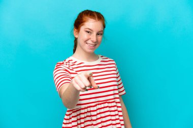 Mavi arka planda izole edilmiş genç, kırmızı bir kadın, mutlu bir ifadeyle ön tarafı işaret ediyor.