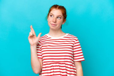 Kırmızımsı genç kadın mavi arka planda izole edilmiş, parmaklarını çapraz yapıp en iyisini diliyor.