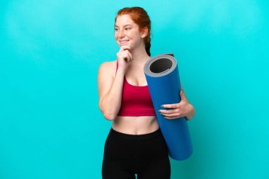 Genç spor kırmızısı kadın yoga derslerine giderken mavi arka planda bir minder tutuyor ve yan tarafa bakıyor ve gülümsüyor.