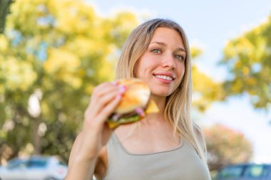 Genç sarışın bir kadın açık havada elinde mutlu bir ifadeyle bir burger tutuyor.