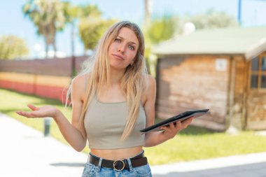 Genç sarışın kadın açık havada bir tablet tutuyor. Omuzlarını kaldırırken şüpheyle hareket ediyor.