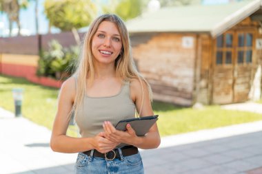Açık havada elinde tablet olan genç sarışın kadın gülümsüyor.