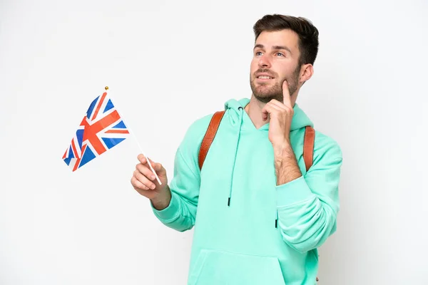 白人を背景に孤立したイギリスの旗を掲げている若い学生の白人男性がアイデアを考えながら見上げ — ストック写真