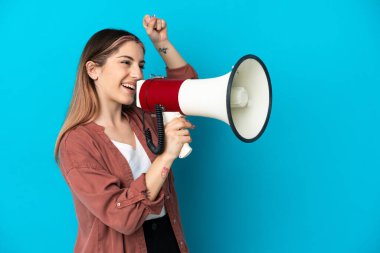 Mavi arka planda izole edilmiş genç beyaz bir kadın megafondan bir şey duyurmak için bağırıyor.