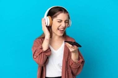 Genç beyaz kadın mavi arka planda izole edilmiş. Cep telefonuyla müzik dinliyor ve şarkı söylüyor.