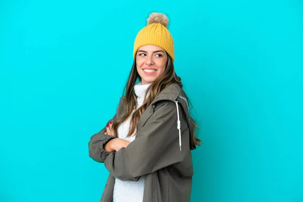Young Italian woman wearing winter jacket and hat isolated on blue background with arms crossed and happy