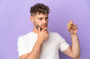 Young caucasian man holding a Bitcoin isolated on purple background having doubts and thinking