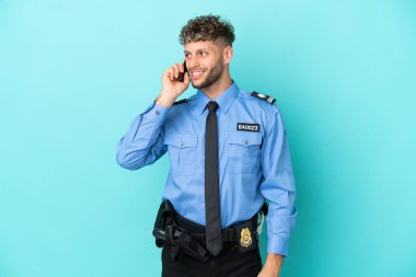 Young police blonde man isolated white on blue background keeping a conversation with the mobile phone with someone
