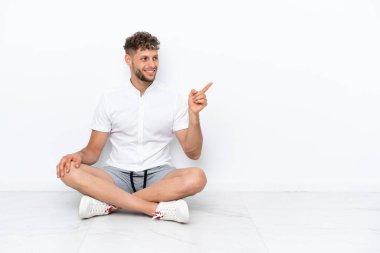 Young blonde man sitting on the floor isolated on white background pointing finger to the side and presenting a product