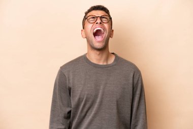 Genç Brezilyalı adam bej arka planda izole edilmiş ve ağzı açık bağırıyor.