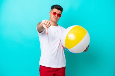 Young caucasian man holding a beach ball isolated on blue background showing thumb down with negative expression