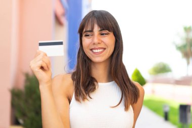 Açık havada elinde mutlu bir ifadeyle kredi kartı tutan genç bir kadın.
