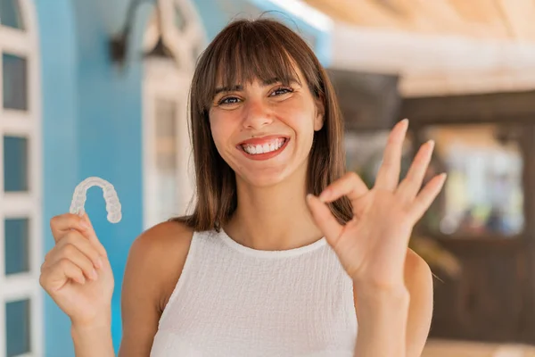 Young woman holding invisible braces at outdoors showing ok sign with fingers